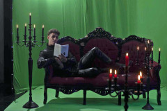 Johannes Lima, zurechtgestylt als düsterer Vampir, liegt auf einem Barocksofa vor einem Green Screen Hintergrund, umgeben von schwarzen Kerzenleuchtern, die die Szene geheimnisvoll illuminieren. Doch statt sich in Blutlust zu verlieren, offenbart er seine intellektuelle Seite: In den Händen hält er das Buch „Vampire!: Schattengewächse der Aufklärung. Über uns aufgeklärte Menschen im Angesicht der Un-Toten“ von Andreas Puff-Trojan, das er mit sichtbarer Neugier liest. So wird der Vampir hier nicht nur als Verführer, sondern auch als literaturaffiner Denker und Bildungshungriger inszeniert – ein Wesen zwischen Sinnlichkeit und Geist.